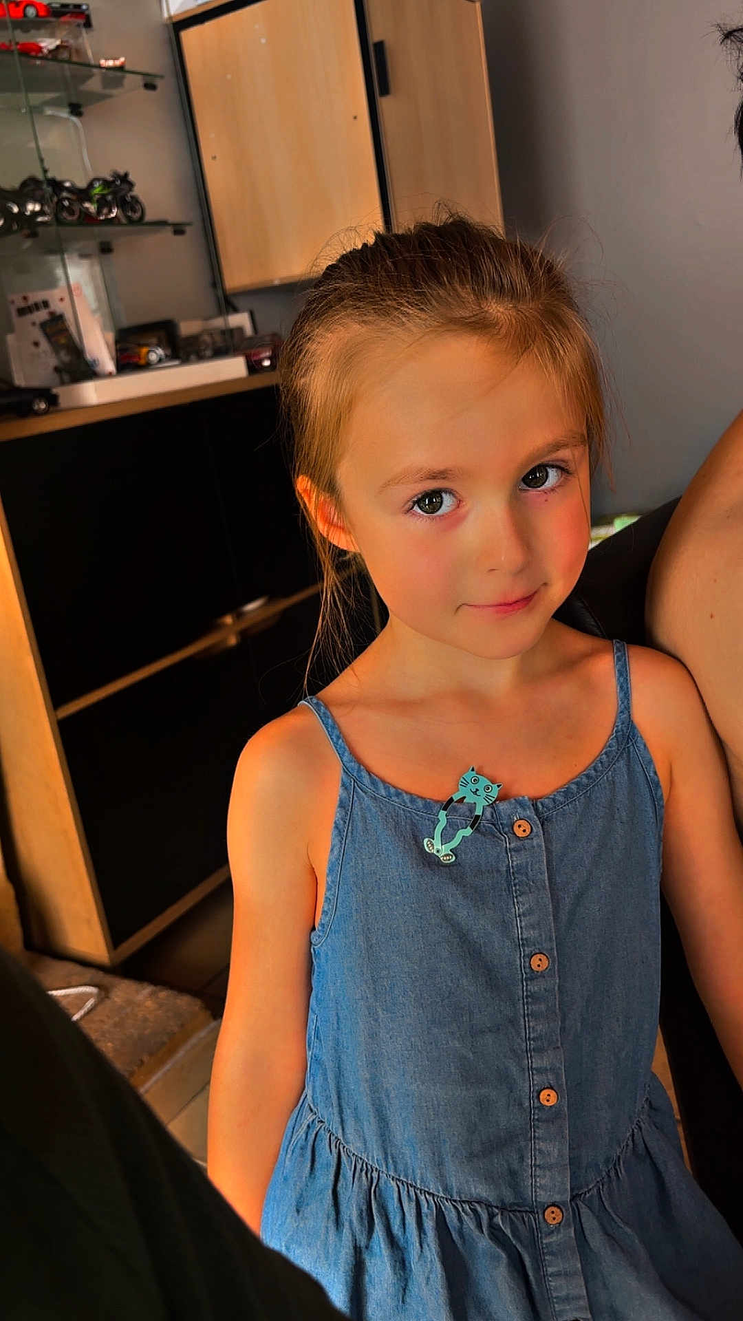 Lya participe au concours pour gagner de l'argent avec cette photo : child, girl, denim_dress, cat_pin, indoor, smiling, portrait, young, person, cabinet, furniture, shelf, toy_cars, hair_tied_back, skin, buttoned_dress, casual_clothing, home, cute, face