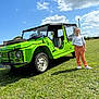 child, car, vintage_car, green_car, grass, outdoor, field, blue_sky, clouds, child_standing, person, casual_clothing, pants, sweater, shoes, daylight, vehicle, transportation, nature, sunny
