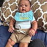 baby, infant, pillow, bed, bib, clothing, shorts, shirt, red_bracelet, hand, face, eyes, mouth, skin, furniture, pattern, home, relaxed, lying_down, cute