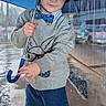 child, umbrella, rain, wet_pavement, gray_sweater, navy_shorts, blue_umbrella, outdoor, rainy_day, curious_expression, footwear, shorts, boy, clothing, street, urban, weather, portrait, standing, daylight