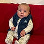 baby, infant, child, couch, red_couch, vest, onesie, clothing, indoor, face, hands, feet, seated, curious, wide_eyes, plush, portrait, cute, cozy, home