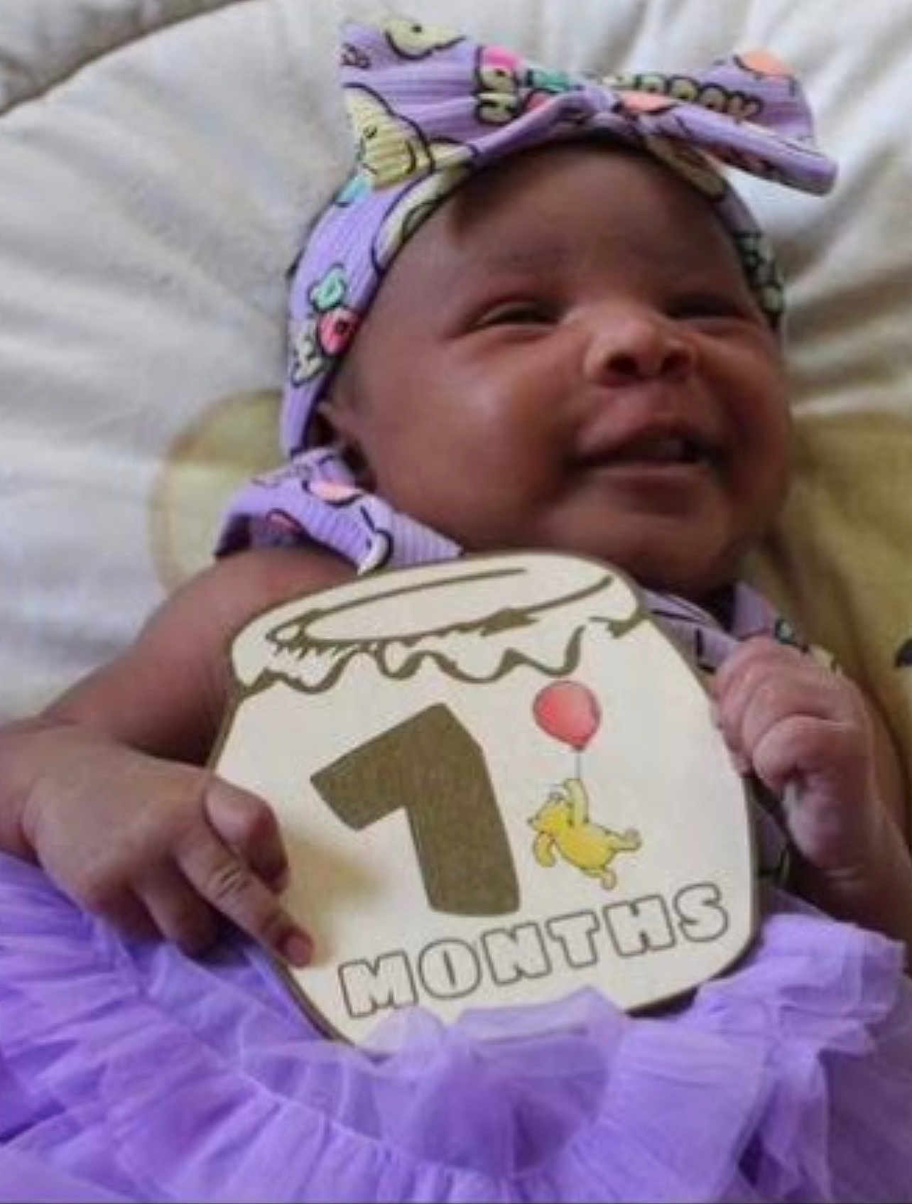 Naomi is registered to the contest to win money with this photo: baby, infant, smiling, purple_outfit, headband, sign, celebration, milestone, month_marker, cute, happy, child, portrait, indoors, cushion, hand, face, person, tutu, soft