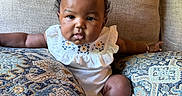 Novèn is registered to the contest to win money with this photo: arm, baby, child, chubby_cheeks, couch, curly_hair, cute, embroidery, face, foot, furniture, hand, indoors, infant, patterned_pillow, pillow, portrait, sitting, skin, white_outfit