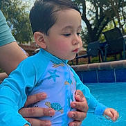 Thiago is registered to the contest to win money with this photo: child, toddler, baby, swimming, pool, water, outdoor, summer, portrait, side_profile, wet_hair, blue_clothing, rash_guard, sea_animals, turtle_pattern, starfish_pattern, adult_hands, family, backyard, sunlight