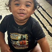 Zion is registered to the contest to win money with this photo: toddler, child, smiling, curly_hair, black_tshirt, diaper, sitting, rug, indoor, happy, cute, face, person, baby, young_child, portrait, expression, clothing, floor, home