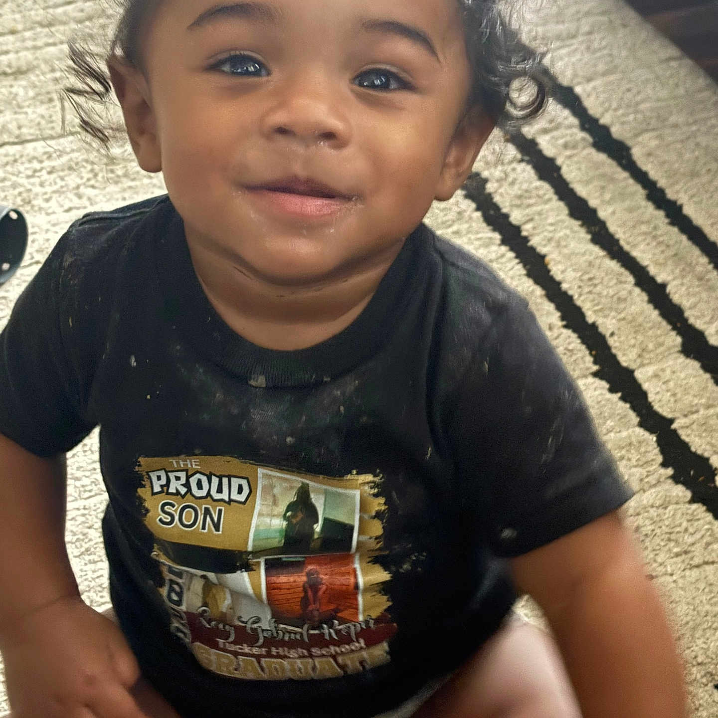 Zion is registered to the contest to win money with this photo: toddler, child, smiling, curly_hair, black_tshirt, diaper, sitting, rug, indoor, happy, cute, face, person, baby, young_child, portrait, expression, clothing, floor, home