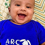 Yashvik is registered to the contest to win money with this photo: baby, smiling, blue_shirt, infant, happy, face, child, pediatrics, text_on_clothing, patterned_background, closeup, cute, portrait, laying_down, young_child, cheerful, indoors, head, person, apparel