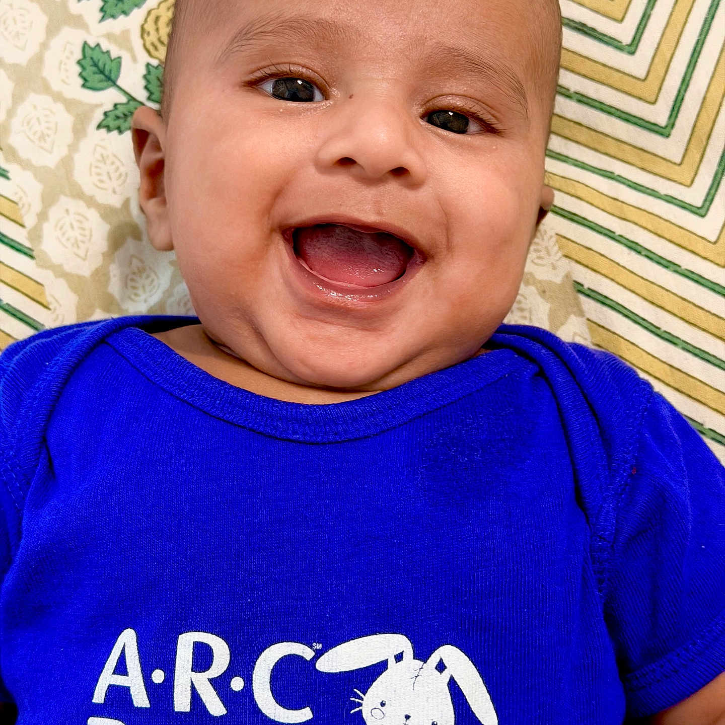 Yashvik is registered to the contest to win money with this photo: apparel, baby, blue_shirt, cheerful, child, closeup, cute, face, happy, head, indoors, infant, laying_down, patterned_background, pediatrics, person, portrait, smiling, text_on_clothing, young_child