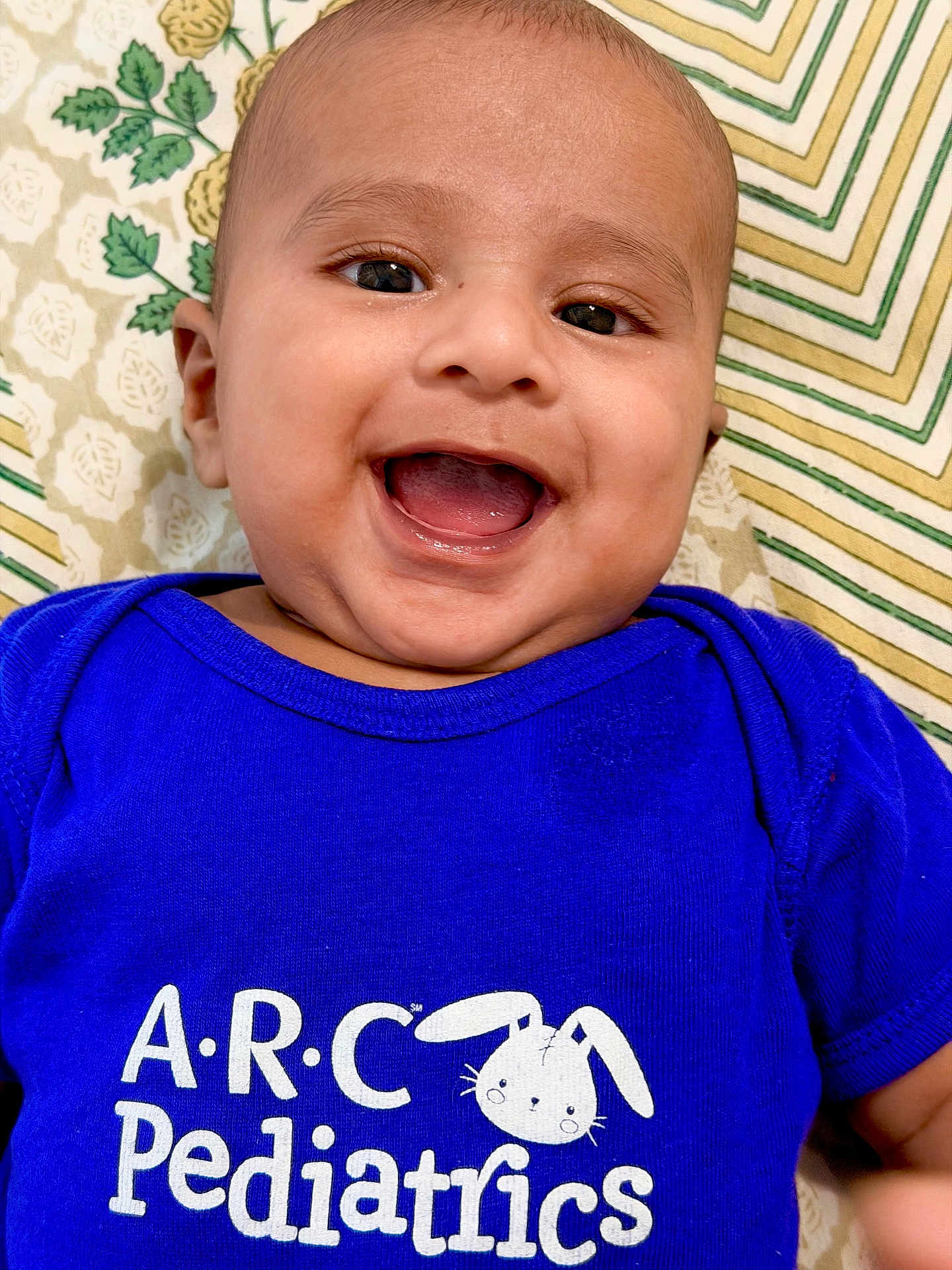Yashvik is registered to the contest to win money with this photo: baby, smiling, blue_shirt, infant, happy, face, child, pediatrics, text_on_clothing, patterned_background, closeup, cute, portrait, laying_down, young_child, cheerful, indoors, head, person, apparel