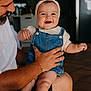 baby, child, smile, denim_overalls, headband, adult, indoor, happy, person, holding, face, legs, hand, beard, casual_clothing, kitchen, refrigerator, warm_lighting, closeup, family