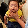 baby, child, person, hand, yellow_shirt, blue_pants, indoor, chair, table, hair, face, eyes, expression, holding, adult_hand, sitting, clothing, skin, portrait, cute