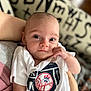 baby, infant, person, face, eyes, hand, shirt, clothing, white, logo, baseball, yankees, holding, blurred_background, indoor, cute, young_child, portrait, expression, soft_light