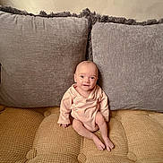 Nova is registered to the contest to win money with this photo: baby, infant, couch, pillow, gray_pillow, beige_couch, onesie, clothing, indoor, smile, face, person, sitting, child, young, cute, home, soft_texture, comfortable, relaxed