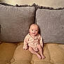 baby, infant, couch, pillow, gray_pillow, beige_couch, onesie, clothing, indoor, smile, face, person, sitting, child, young, cute, home, soft_texture, comfortable, relaxed