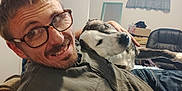 Colby is registered to the contest to win money with this photo: man, glasses, dog, smile, indoor, window, curtain, couch, shirt, hand, relaxing, pet, home, furniture, casual, portrait, happy, beard, brown_hair, resting