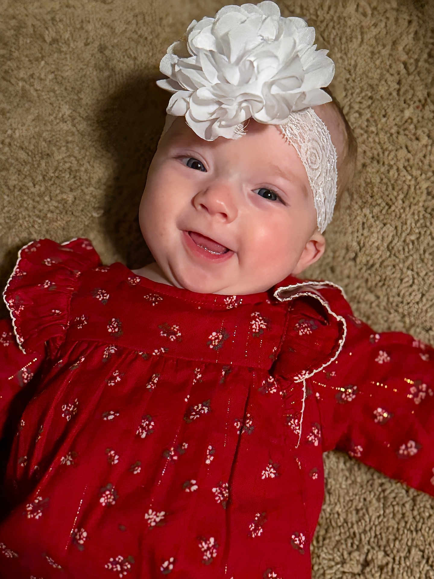 Ellie is registered to the contest to win money with this photo: baby, infant, smile, headband, flower, red_dress, carpet, portrait, child, cute, happy, face, young, clothing, laying_down, indoors, closeup, person, head, skin