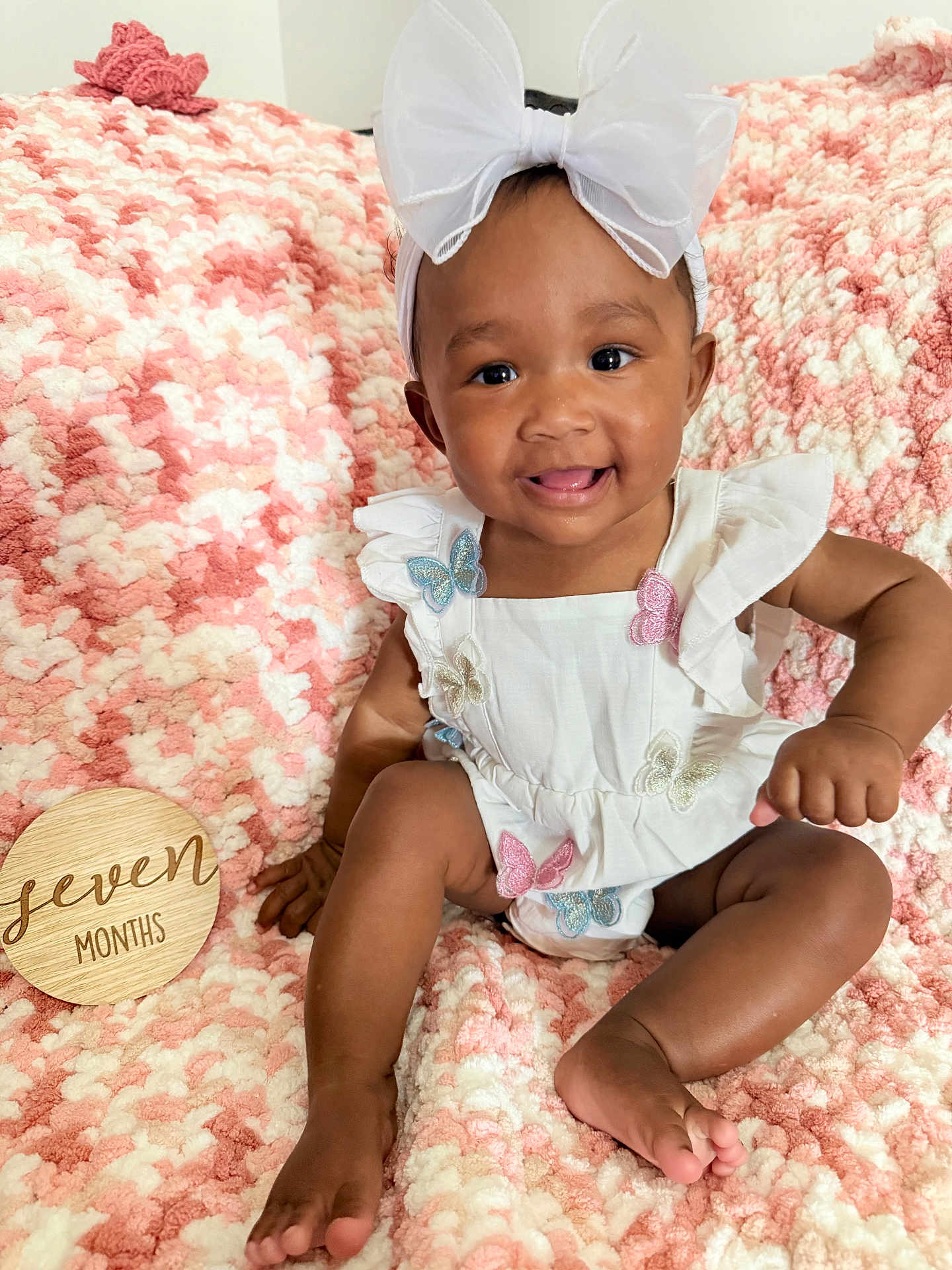 Melodie joined the competition — help win amazing prizes! baby, child, infant, smiling, white_bow, butterfly, headband, white_outfit, pink_blanket, textured_blanket, wooden_sign, seven_months, happy, cute, sitting, tongue_out, indoors, portrait, soft_texture, young_child
