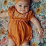 baby, infant, child, portrait, lying_down, smiling, orange_outfit, romper, hat, topknot, socks, floral_blanket, bed, indoor, close_up, face, eyes, arms, fashion, cute