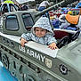 toddler, child, hooded_jacket, amusement_park, mini_tank, us_army, ride, smile, outdoor, playful, carnival, colorful, toy_vehicle, fun, face, person, clothing, cloudy_sky, hand, park