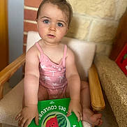 Gabrielle participe au concours pour gagner de l'argent avec cette photo : toddler, child, blue_eyes, pink_outfit, chair, book, watermelon, brick_wall, stone_wall, indoors, curious, sitting, hand, foot, wooden_armrest, reading, portrait, person, baby, cute