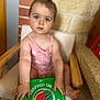 toddler, child, blue_eyes, pink_outfit, chair, book, watermelon, brick_wall, stone_wall, indoors, curious, sitting, hand, foot, wooden_armrest, reading, portrait, person, baby, cute