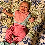 baby, infant, smiling, money, dollar_bills, pink_clothing, bib, blue_socks, floral_bedspread, happy, lying_down, indoors, person, currency, cute, small_child, face, hand, footwear, background