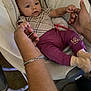 adult_hands, baby, baby_seat, blanket, carpet, child, clothing, comfort, cushion, cute, footwear, hands, holding_hands, indoor, infant, person, purple_pants, relaxation, socks, toy
