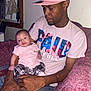 Samantha is registered to the contest to win money with this photo: man, baby, child, indoor, couch, pink, cap, shirt, plaid, pants, holding, seated, person, portrait, family, casual, home, comfort, relaxed, love