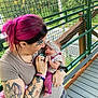 baby, woman, tattoos, pink_hair, holding, outdoor, balcony, metal_railings, plaid_pants, headband, infant, affection, person, casual_clothing, nature, wooden_floor, smile, bracelets, rings, closeup
