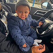 Adonis is registered to the contest to win money with this photo: adult, car_interior, child, cozy, dinosaur_pattern, hand, happy, hat, jacket, person, playful, seat, smiling, snowsuit, steering_wheel, sunlight, toddler, vehicle, window, winter_clothing