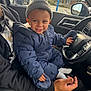 adult, car_interior, child, cozy, dinosaur_pattern, hand, happy, hat, jacket, person, playful, seat, smiling, snowsuit, steering_wheel, sunlight, toddler, vehicle, window, winter_clothing
