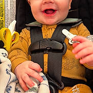Ryker is registered to the contest to win money with this photo: baby, infant, smiling, car_seat, harness, blanket, toy_banana, hand, face, blue_eyes, onesie, cheek, closeup, portrait, happy, cute, seat_buckle, soft_texture, toddler, indoor