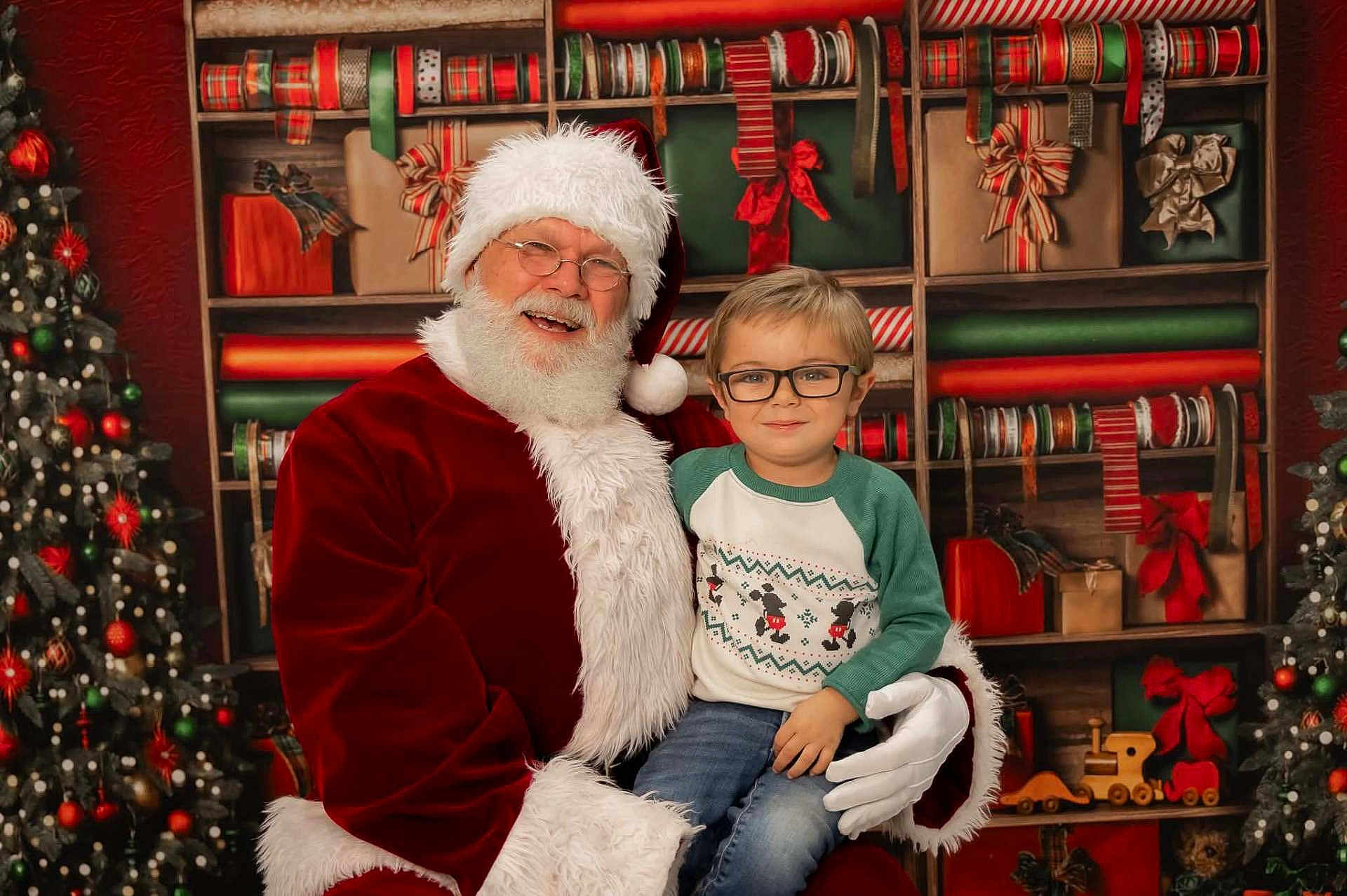 Niko is registered to the contest to win money with this photo: santa_claus, child, glasses, christmas, holiday, gift_wrap, tree, decorations, smile, festive, red_suit, white_beard, toy_train, shelf, young_boy, indoor, portrait, happy, winter_clothing, celebration
