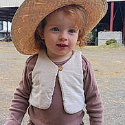 Fréya participe au concours pour gagner de l'argent avec cette photo : barn, belt, blue_eyes, brown_clothing, building, child, curly_hair, cute, daylight, hat, outdoor, pants, person, portrait, sky, smile, standing, straw_hat, toddler, vest
