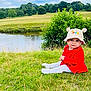 baby, child, grass, river, hat, red_clothing, outdoor, nature, trees, sky, clouds, field, greenery, cute, sitting, portrait, person, daylight, scenery, smile