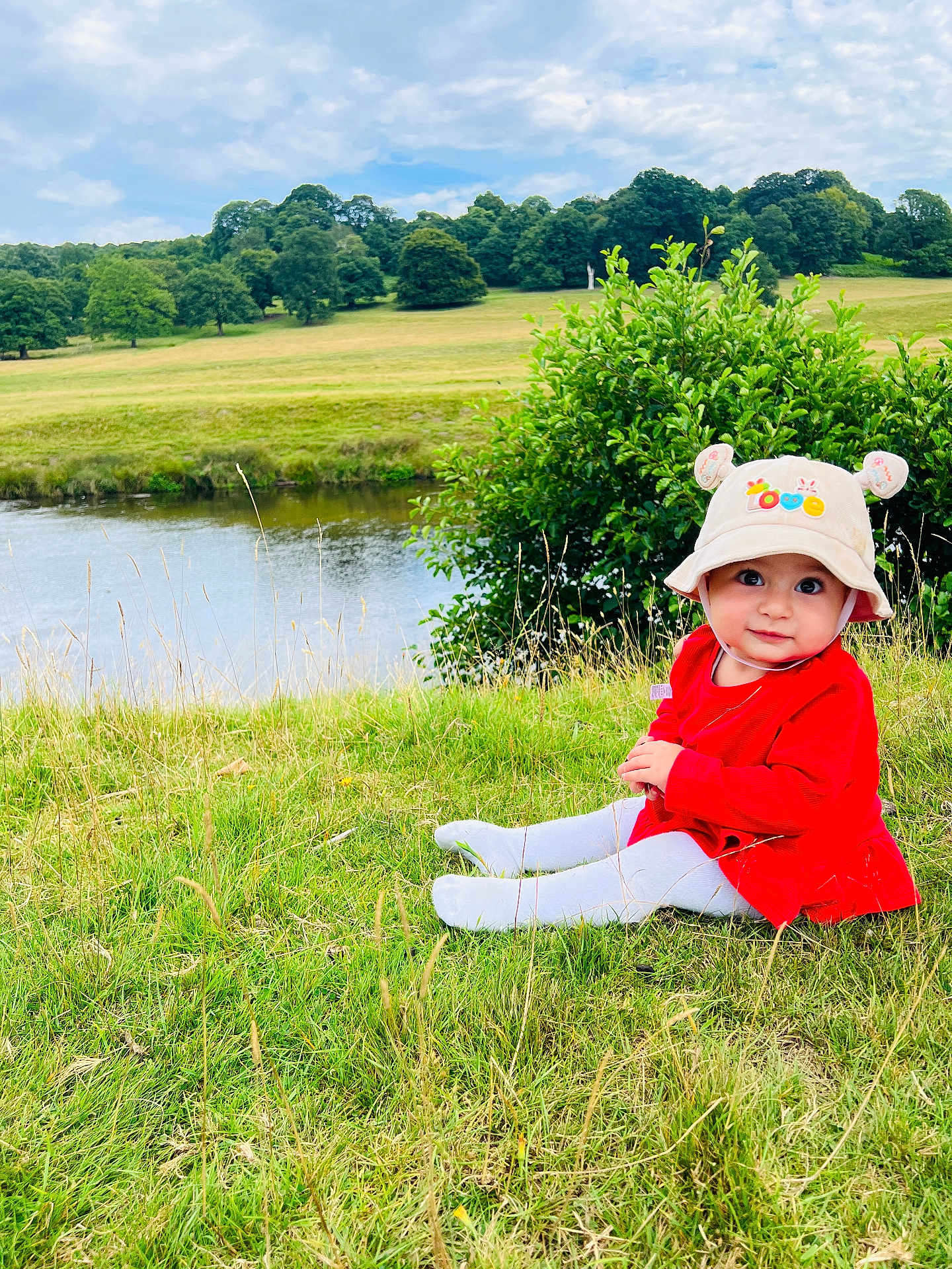 Alin joined the competition — help win amazing prizes! baby, child, grass, river, hat, red_clothing, outdoor, nature, trees, sky, clouds, field, greenery, cute, sitting, portrait, person, daylight, scenery, smile