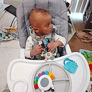 Terrence is registered to the contest to win money with this photo: baby, blanket, carpet, clothing, colorful_toy, couch, face, feeding_tray, floor, hands, harness, head, high_chair, infant, living_room, looking_sideways, play_mat, tablet, toy, vertical_blinds