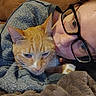 person, cat, orange_tabby, glasses, blanket, couch, indoor, close_up, snuggling, cozy, relaxed, pet, feline, warm, casual, human_face, animal, comfort, resting, eyewear