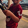 child, toddler, smiling, red_shirt, outdoor, parking_lot, car, concrete, person, curly_hair, holding, side_view, urban, daylight, casual_clothing, screenshot, social_media, video, user_interface, happy