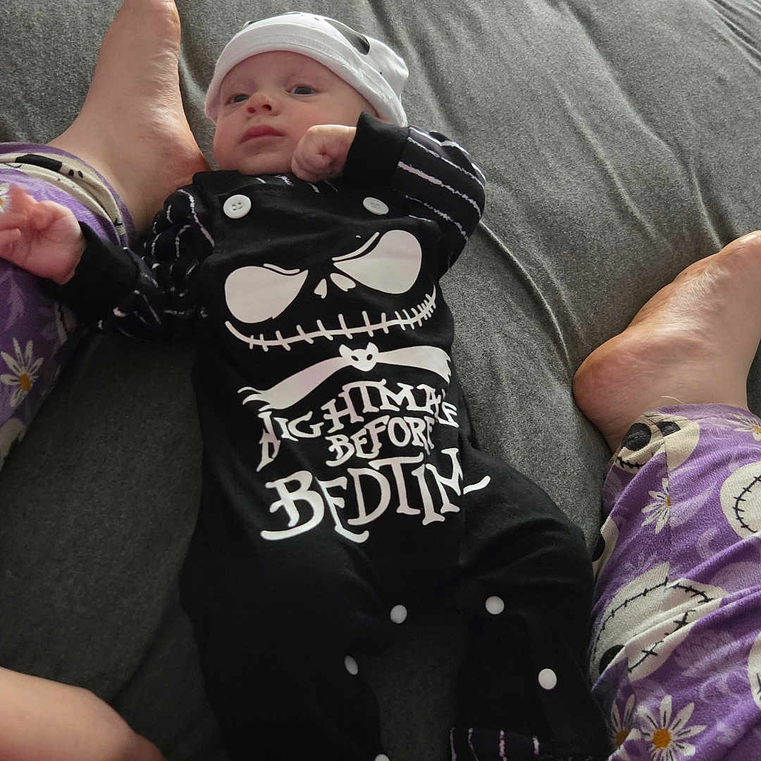 Andrew joined the competition — help win amazing prizes! baby, infant, child, newborn, face, portrait, lying_down, bed, bedroom, skeleton_costume, black_clothing, hat, onesie, pajamas, adult_feet, family, indoor, cozy, funny, photograph