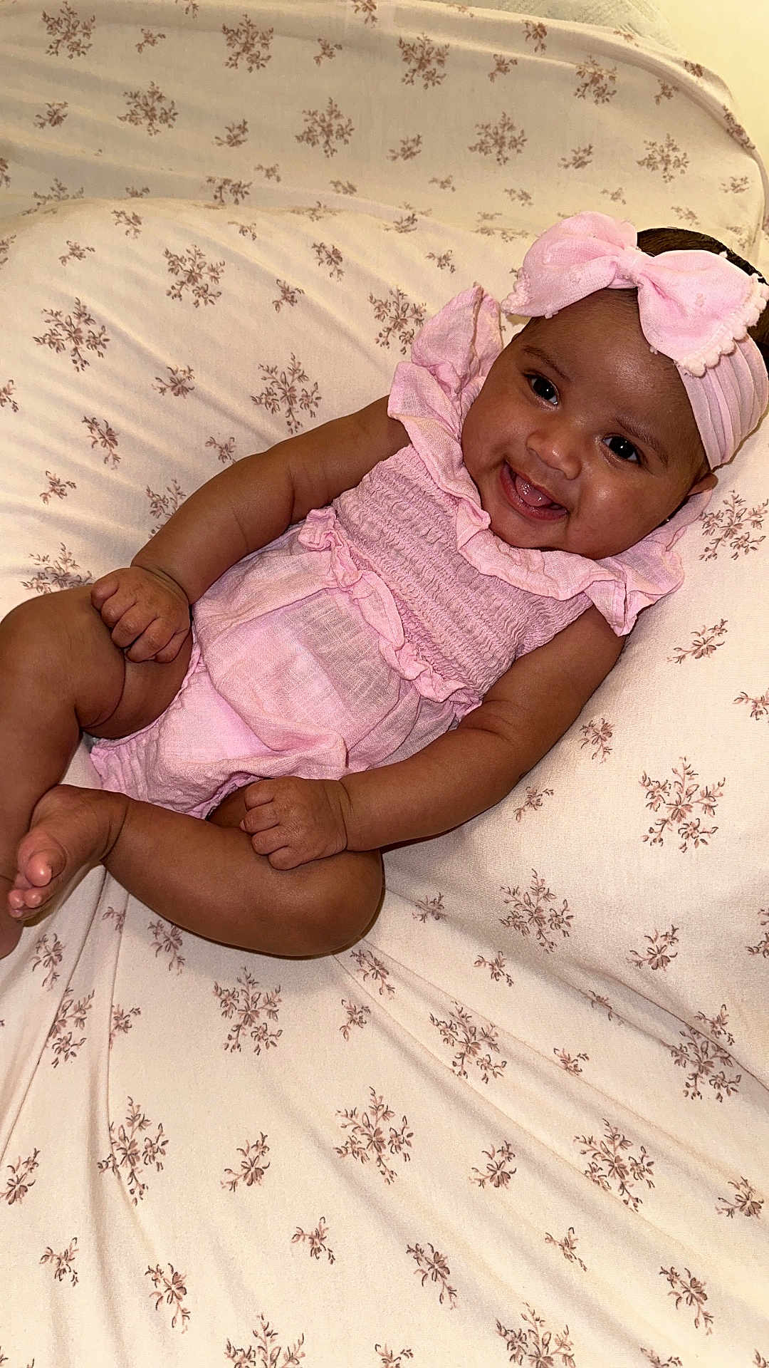 Riahleigh is registered to the contest to win money with this photo: baby, infant, smiling, pink_outfit, headband, lying_down, floral_pattern, bedspread, cute, child, happy, skin, face, hands, feet, indoors, portrait, toddler, young_child, sleepwear