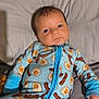 baby, infant, onesie, blue_clothing, bed, pillow, sleepwear, face, newborn, cute, patterned_clothing, child, person, lying_down, soft_fabric, indoors, young_child, expression, sleepy, cozy