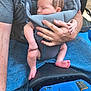 baby, baby_carrier, adult, hand, gray_tshirt, blue_jeans, blue_vehicle, seat, sleeping, skin, person, outdoor, foot, arm, clothing, hand_holding, transport, comfort, rest, closeup