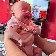 Ophelia is registered to the contest to win money with this photo: baby, infant, smile, laughing, pink_onesie, arms, hands, adult_hands, red_wall, television, living_room, carpet, furniture, person, happy, indoor, closeup, cute, child, home