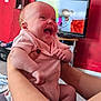 adult_hands, arms, baby, carpet, child, closeup, cute, furniture, hands, happy, home, indoor, infant, laughing, living_room, person, pink_onesie, red_wall, smile, television
