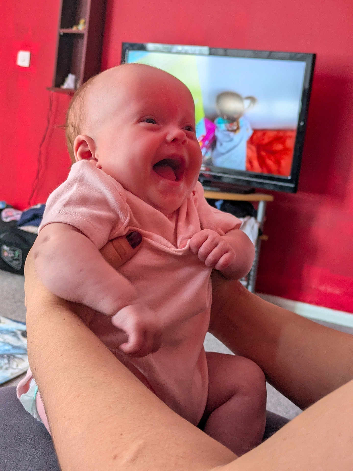 Ophelia is registered to the contest to win money with this photo: baby, infant, smile, laughing, pink_onesie, arms, hands, adult_hands, red_wall, television, living_room, carpet, furniture, person, happy, indoor, closeup, cute, child, home