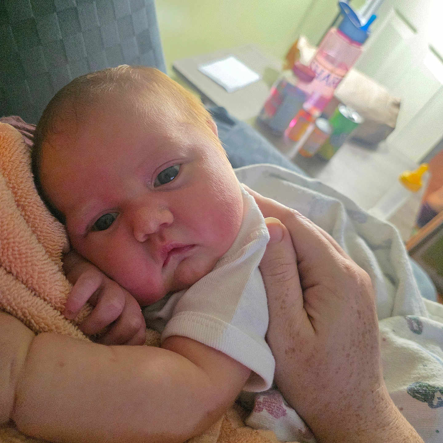 Colbee is registered to the contest to win money with this photo: baby, infant, hand, towel, resting, closeup, indoor, person, freckles, skin, table, water_bottle, cans, background, soft, comfort, home, white_clothing, face, expression