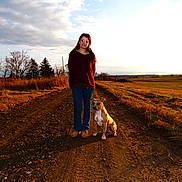 Nugget is registered to the contest to win money with this photo: woman, dog, dirt_road, field, sunset, trees, grass, outdoor, pet, casual_clothing, glasses, landscape, nature, sky, clouds, rural, walking, brown_dog, jeans, sweater