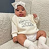 baby, infant, child, white_sweater, headband, bow, sitting, fluffy_chair, white_socks, cute, tongue_out, knitted_clothing, cozy, indoor, portrait, smiling, comfortable, soft_texture, young_child, adorable