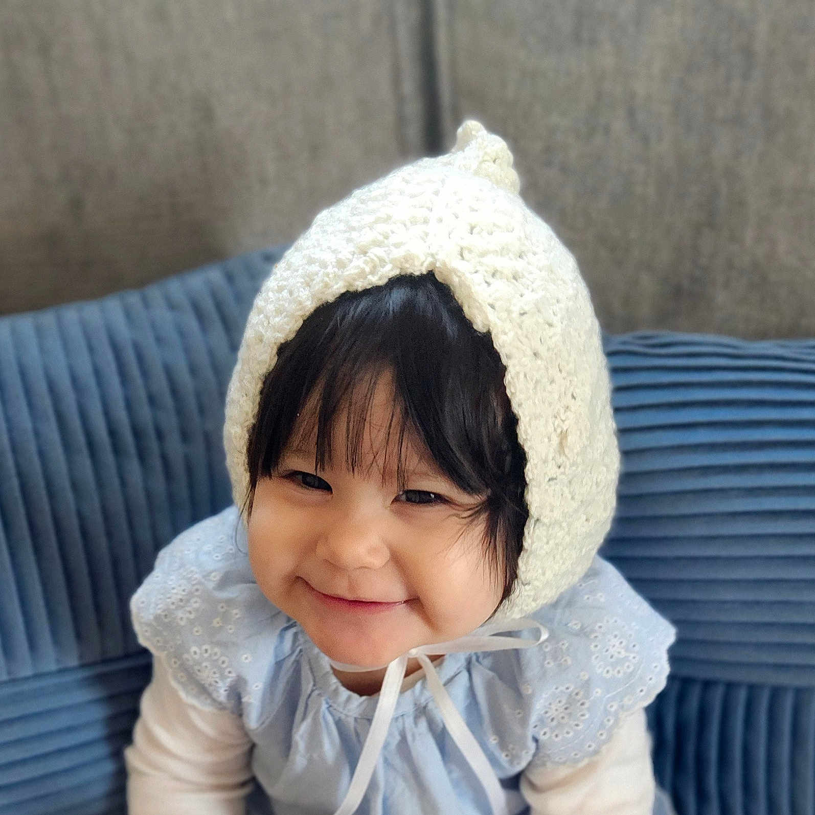 Yael is registered to the contest to win money with this photo: baby, blue_couch, child, closeup, clothing, cute, eyes, fabric, face, happy, headwear, homedecor, indoors, knitted_bonnet, person, portrait, sitting, smile, soft_lighting, toddler